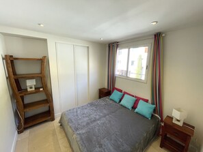 A bedroom with a bed, a wooden shelf unit, a window with curtains, and a bedside table.