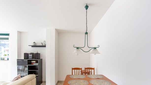 Un espace repas comprenant une table et des chaises en bois, un lustre, ainsi qu’un coin cuisine équipé d’appareils électroménagers et d’un réfrigérateur.