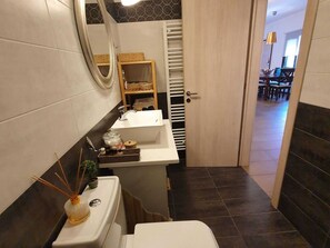 Une petite salle de bain comprenant des toilettes, un lavabo et un miroir. On y trouve un porte-serviettes et une petite étagère avec des articles de toilette. Le sol est carrelé et les murs sont recouverts d’un mélange de carreaux blancs et foncés.