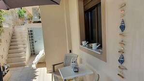 A small outdoor patio with a white table and chairs, a window with decorative items, and a staircase leading to an upper level. - Apartment in Loutra Elenis With Garden (Corinth)