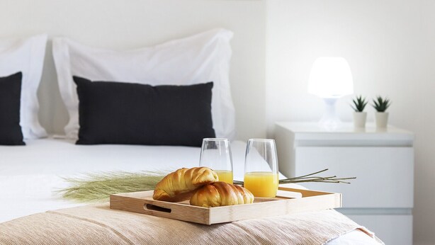 A neatly made bed with white and black bedding, a bedside table with a lamp and two small potted plants, and a tray with croissants and orange juice. - Downtown Palma Suites by Homing (Lisbon)