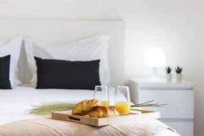 A neatly made bed with white and black bedding, a bedside table with a lamp and two small potted plants, and a tray with croissants and orange juice. - Downtown Palma Suites by Homing (Lisbon)