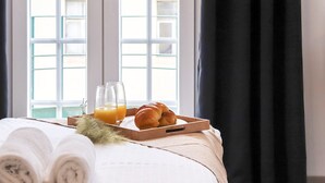 A neatly made bed with rolled towels, a tray with croissants and orange juice glasses, and a window with a view of buildings. - Downtown Palma Suites by Homing (Lisbon)