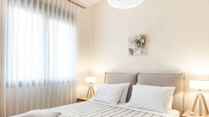 A neatly arranged bedroom with a bed, two bedside tables with lamps, a window with sheer curtains, and a wall-mounted artwork. - Villa in Crete With Stunning Rural Views (Rethymno)