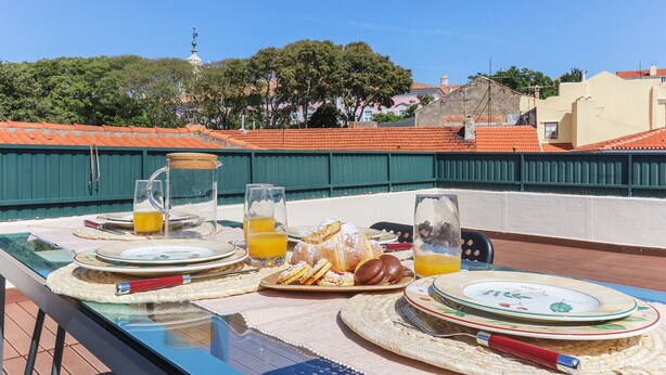 Mesa en una terraza para comer, con platos, vasos y una jarra de jugo de naranja, con vistas a un paisaje urbano con tejados rojos y áreas verdes.