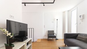 Una sala de estar moderna con un televisor de pantalla plana sobre un soporte blanco, un sofá gris y una silla de mimbre. Hay una planta en maceta sobre el soporte y una mesita en primer plano.