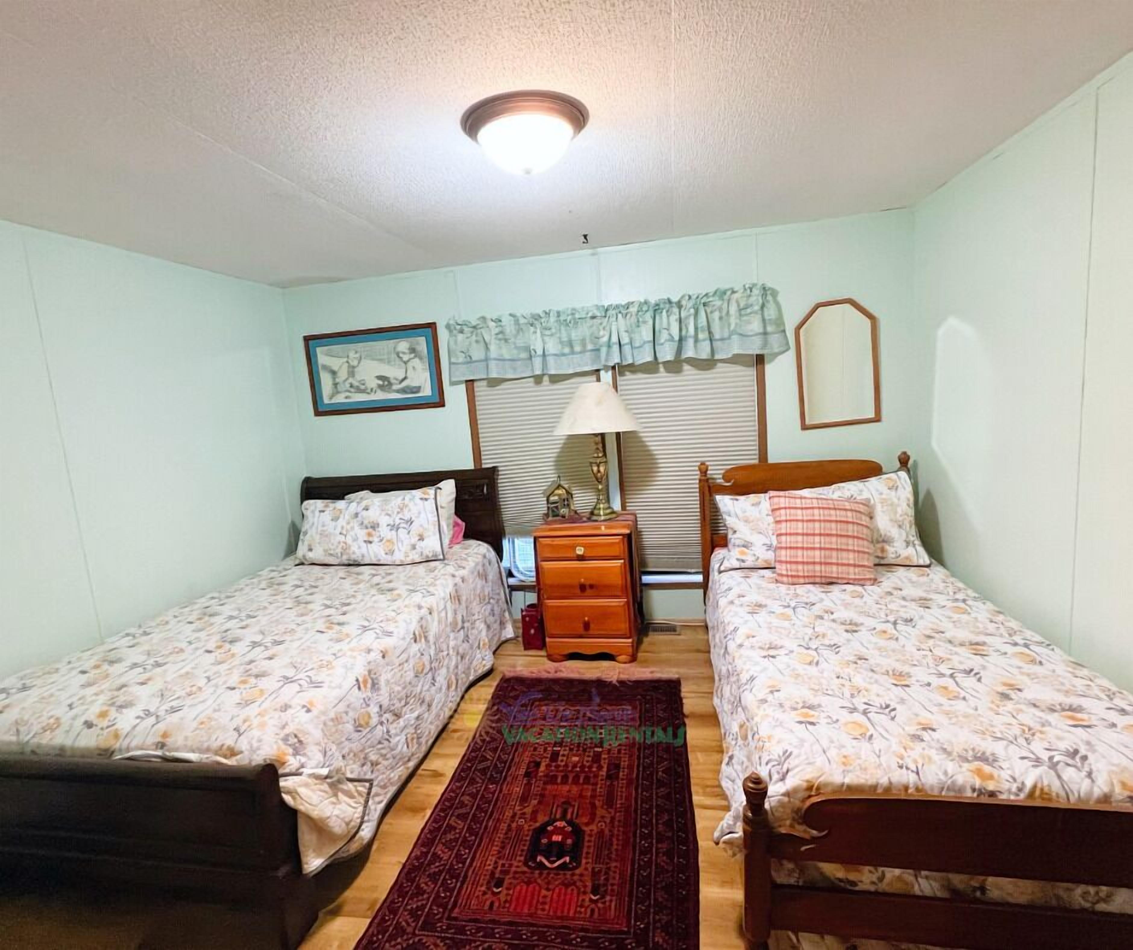 Une chambre avec deux lits simples, une table de chevet avec une lampe, un tableau encadré au mur et une fenêtre avec des stores.