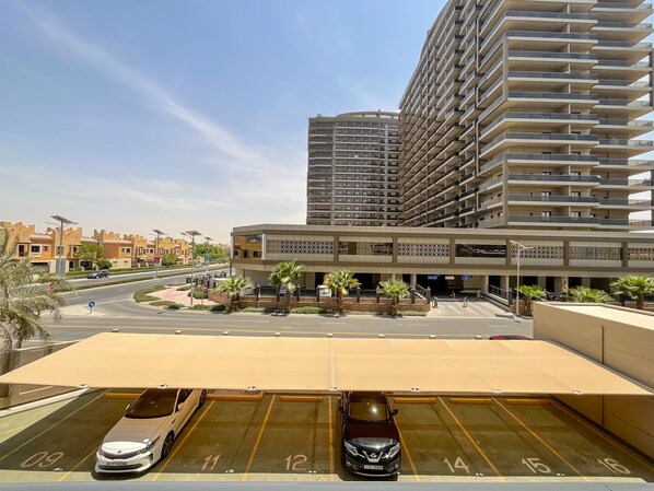 A parking area with multiple cars, a covered section, and a building with balconies in the background.