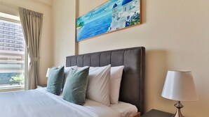 Habitación de hotel con cama, mesita de noche, lámpara, un cuadro de una escena costera y ventana con cortinas.