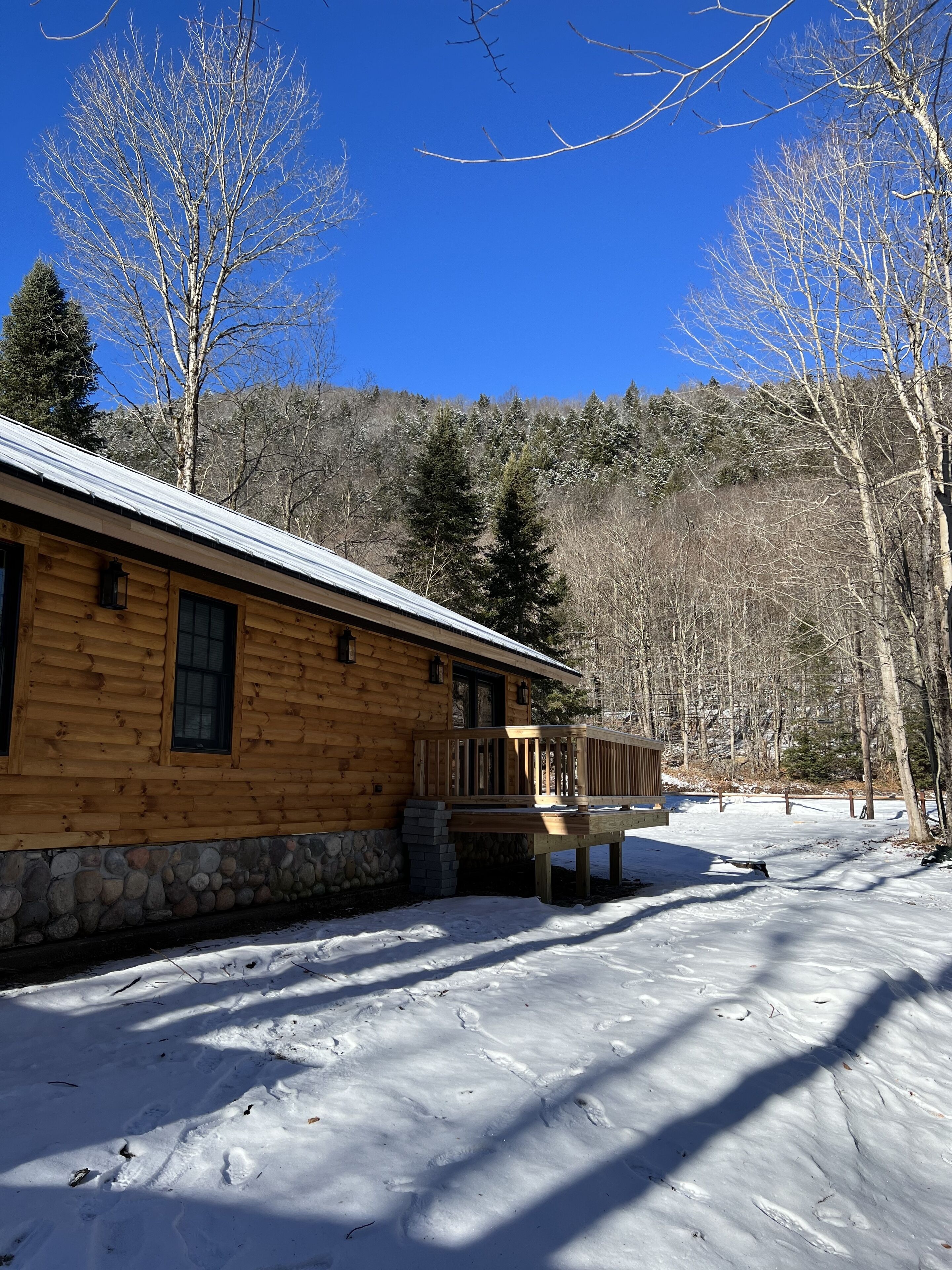 A log cabin with a stone foundation, a wooden deck, and a clear view of the surrounding forest.