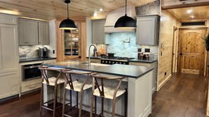 A modern kitchen with a central island, bar stools, a stove, and pendant lighting. - NEW ADIRONDACK LUXURY CABIN WITH YEAR-ROUND HEATED PLUNGE POOL (Old Forge)