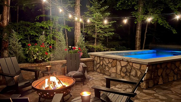 An outdoor patio with a stone wall, a hot tub, and string lights. - NEW ADIRONDACK LUXURY CABIN WITH YEAR-ROUND HEATED PLUNGE POOL (Old Forge)