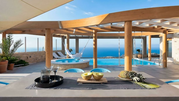 Uma área de piscina ao ar livre com vista para o mar, pérgulas de madeira, espreguiçadeiras e uma mesa com frutas e bandeja de bebidas.