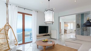 Uma sala de estar moderna com uma televisão grande, um lustre, uma cadeira de vime e vista para o mar.