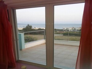 A room with a sliding glass door leading to a balcony with a view of the sea and buildings.