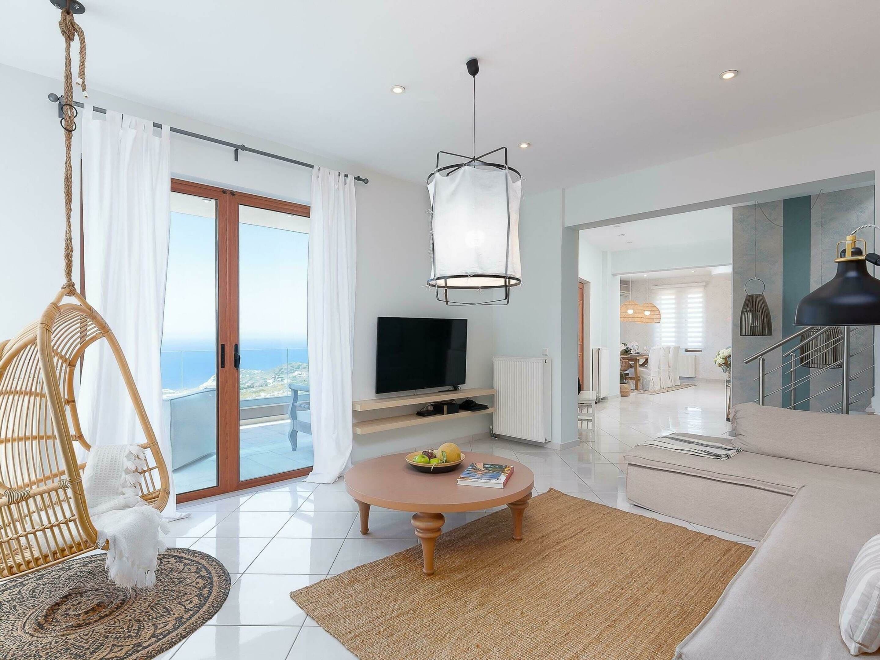 A modern living room with a large TV, a hanging light fixture, a wicker chair, and a view of the ocean.