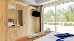 A hotel room with a large bed, a wardrobe with a mirror, an air conditioning unit, a television, and a balcony with a view of trees. - Beautiful Holiday Home With Private Pool (Rhodes)
