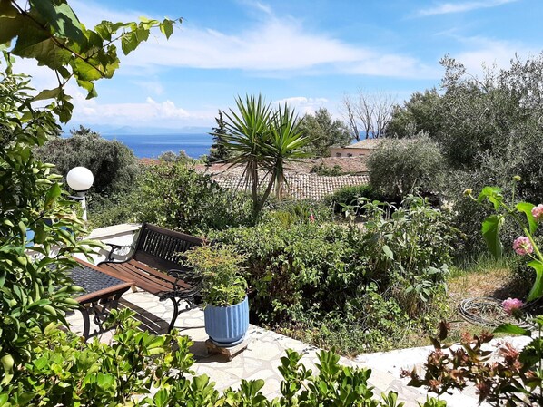 A patio with a bench and potted plants, overlooking a coastal landscape with trees and a view of the sea.