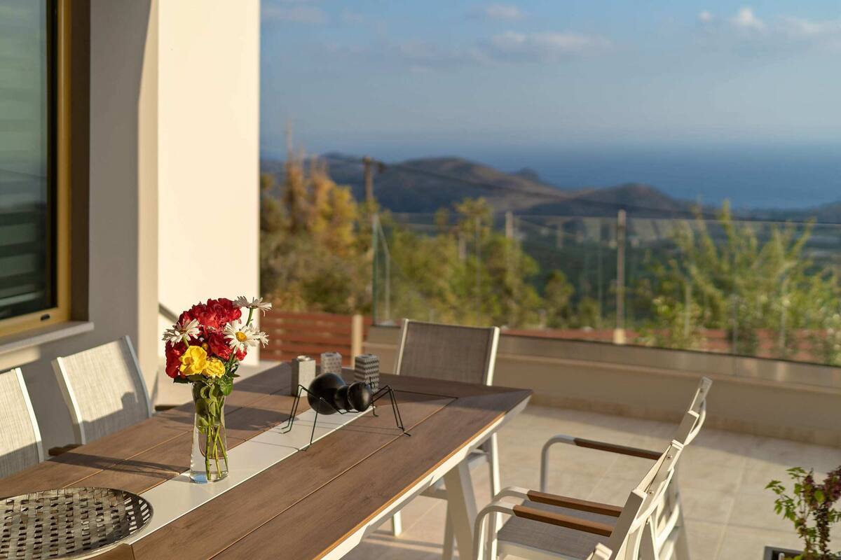 A dining table with chairs, a vase of flowers, and a pair of glasses on a balcony overlooking a scenic landscape.