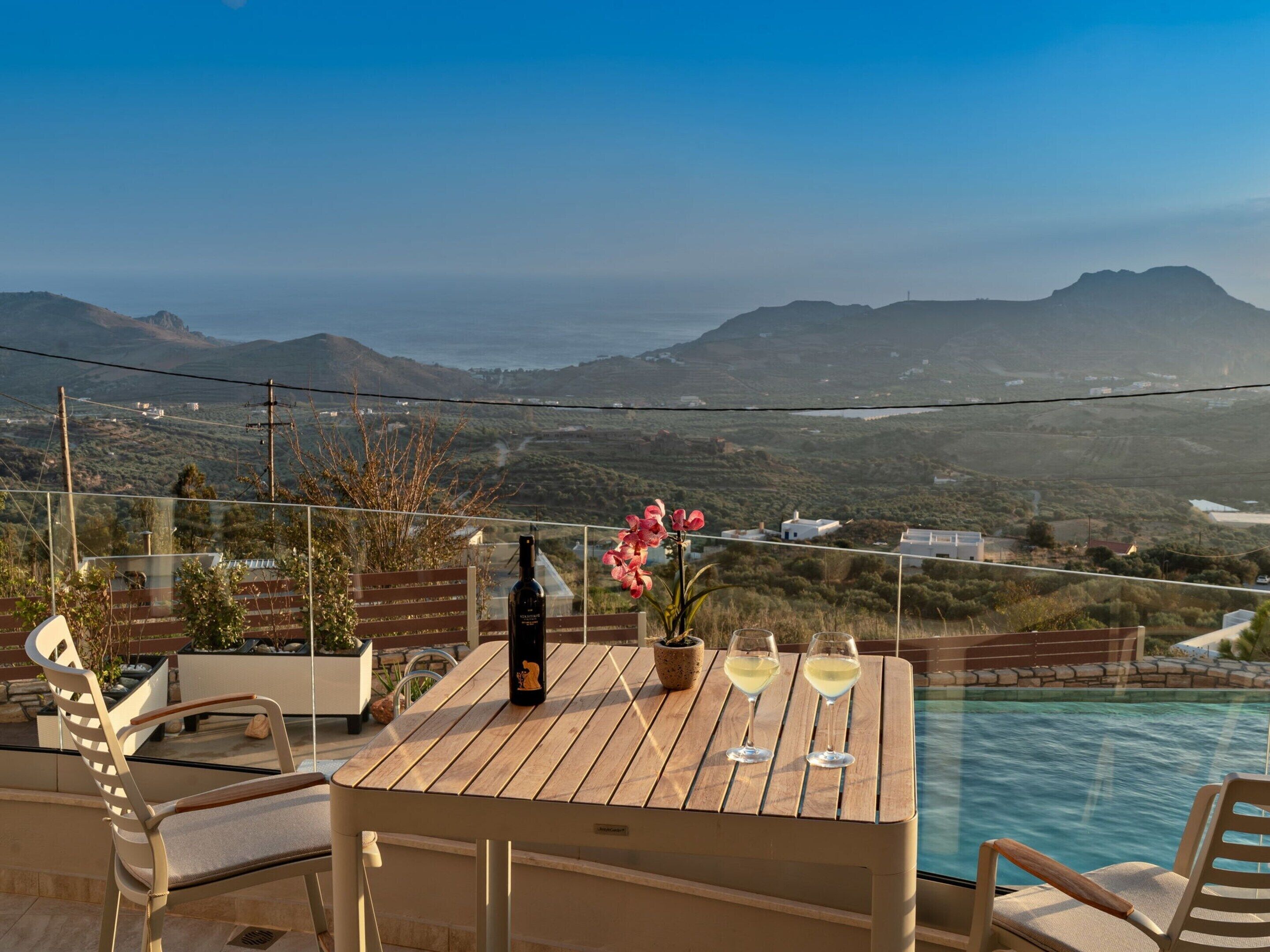 Une table en bois, avec une bouteille et deux verres de vin blanc, donnant sur une piscine et offrant une vue panoramique sur les montagnes et la côte.
