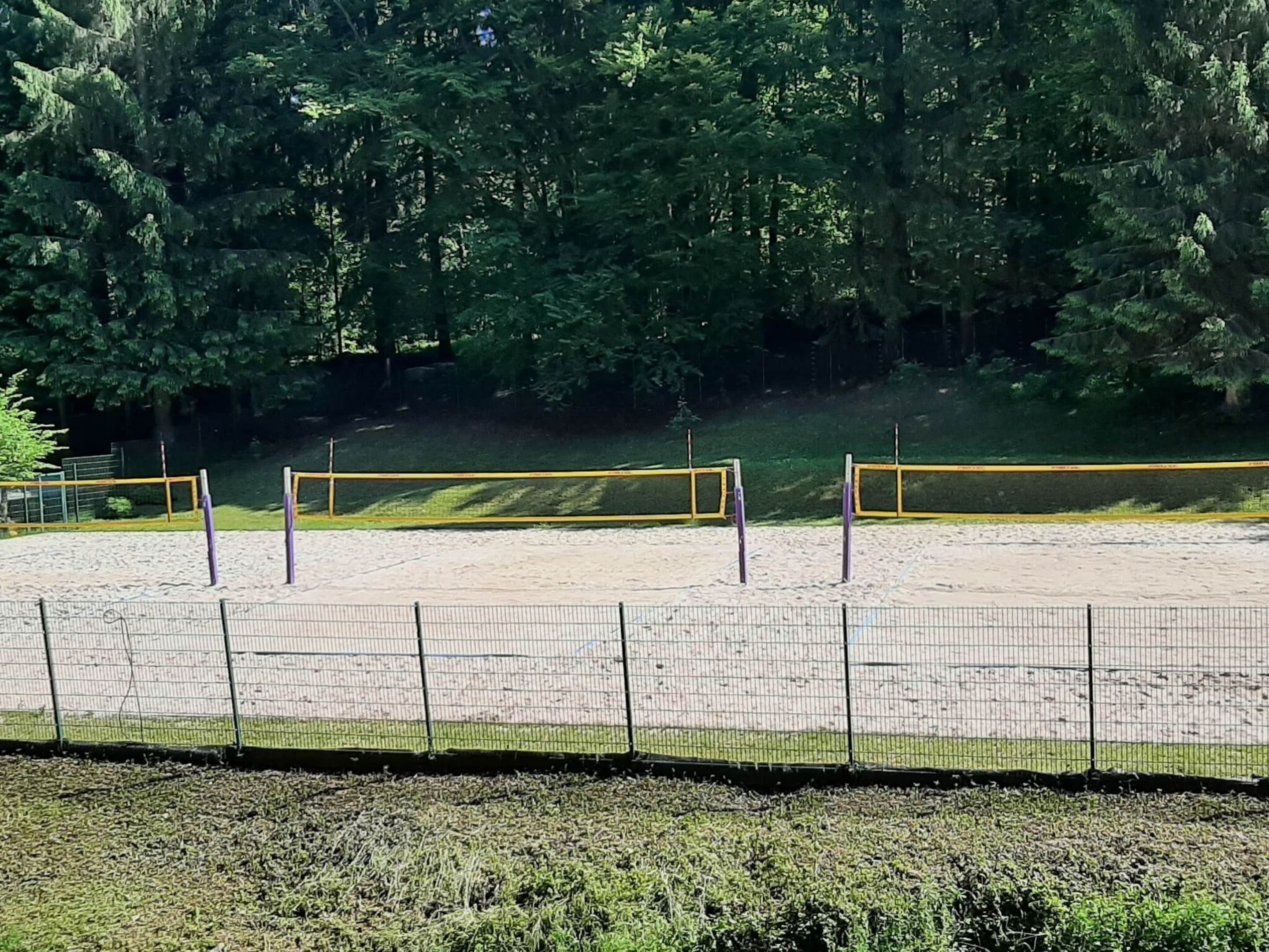 A volleyball court with a net, surrounded by a fence, and a forested area in the background.