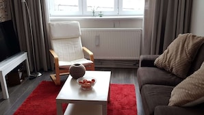 A living room with a white TV stand, a white table with a bowl of fruit, a red rug, a grey sofa with a textured throw pillow, and a window with curtains. - Cosy Holiday Home Near the ski Lift (Grossbreitenbach)