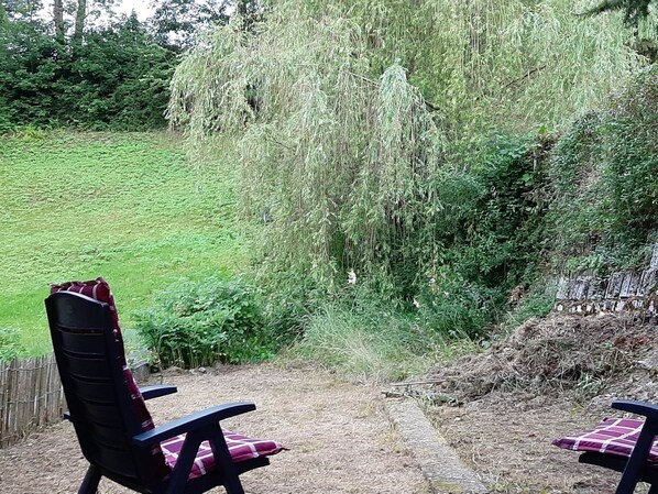 A patio area with two chairs and a table, surrounded by lush greenery and a weeping willow tree.