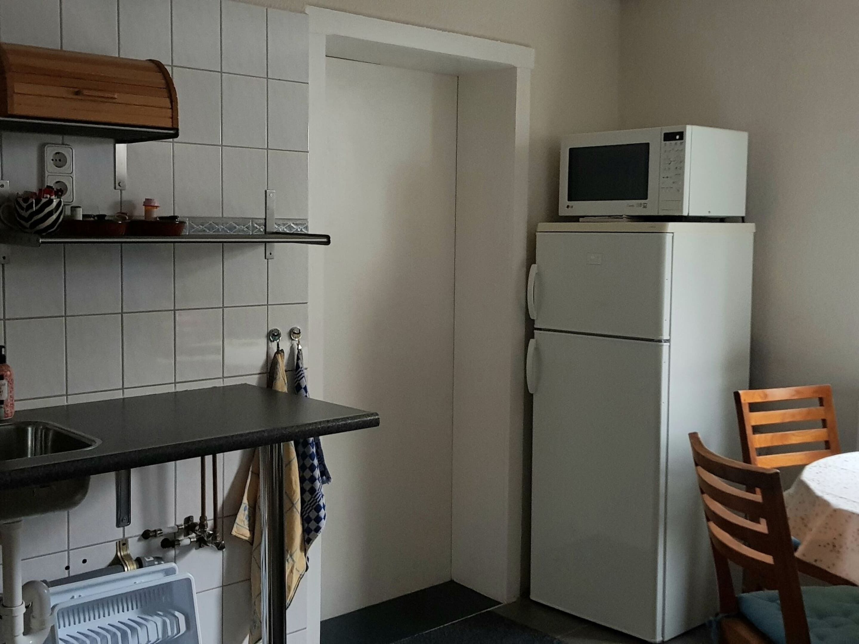 A compact kitchen with white tiled walls, a built-in microwave, a refrigerator, a sink with a faucet, and a wooden chair at a small table.