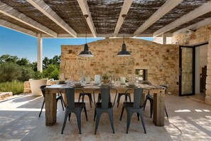 Un espace de restauration en plein air, avec un mur en pierre en arrière-plan, une table en bois dressée pour quatre personnes et deux lampes suspendues.
