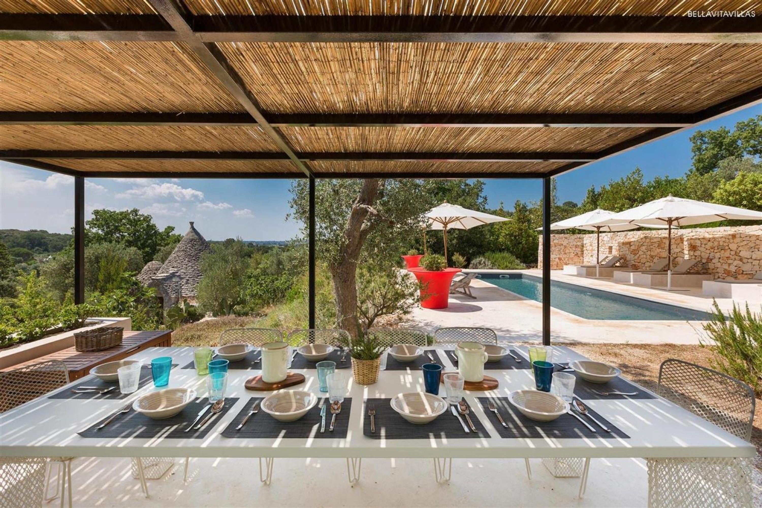 Un espace de restauration en plein air avec une table dressée pour six personnes, offrant une vue sur une piscine et une végétation luxuriante.