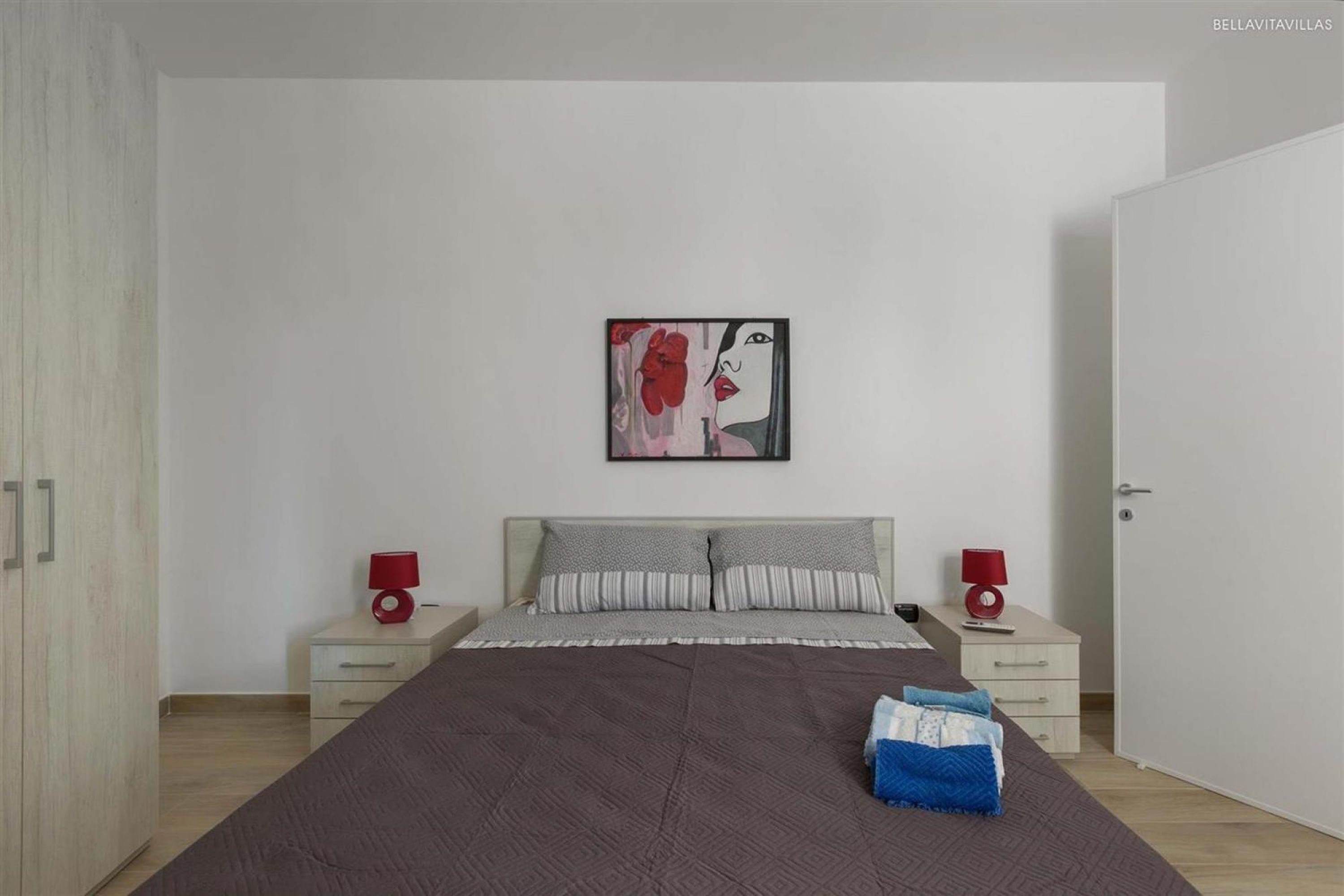 A modern bedroom with a large bed, two bedside tables with lamps, a painting on the wall, and a wardrobe.