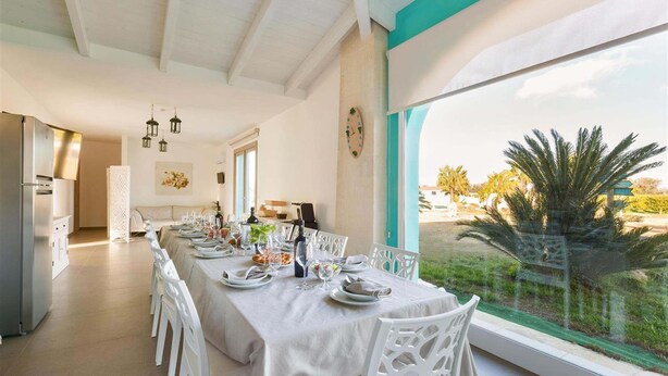 Un comedor con una larga mesa dispuesta para una comida, sillas blancas y vistas al exterior a través de amplios ventanales.