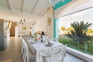 A dining area with a long table set for a meal, white chairs, and a view of the outdoors through large windows. - 2197 Tenuta Donna Caterina (Mesagne)