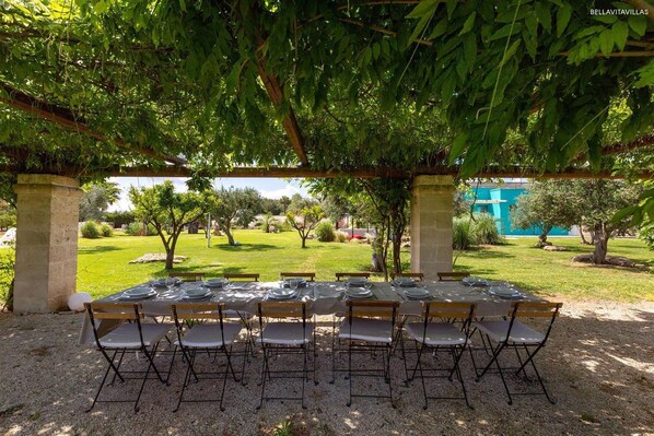 An outdoor dining area with tables set for six, surrounded by trees and a view of a garden and buildings in the background. - 2197 Tenuta Donna Caterina (Mesagne)