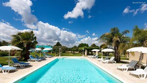 Una piscina con sillas de descanso y sombrillas, rodeada de palmeras y un cielo despejado.