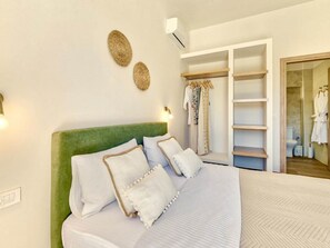 A bedroom with a green upholstered headboard, a bed with white bedding, a wardrobe with hanging clothes, and a bathroom visible through an open door.