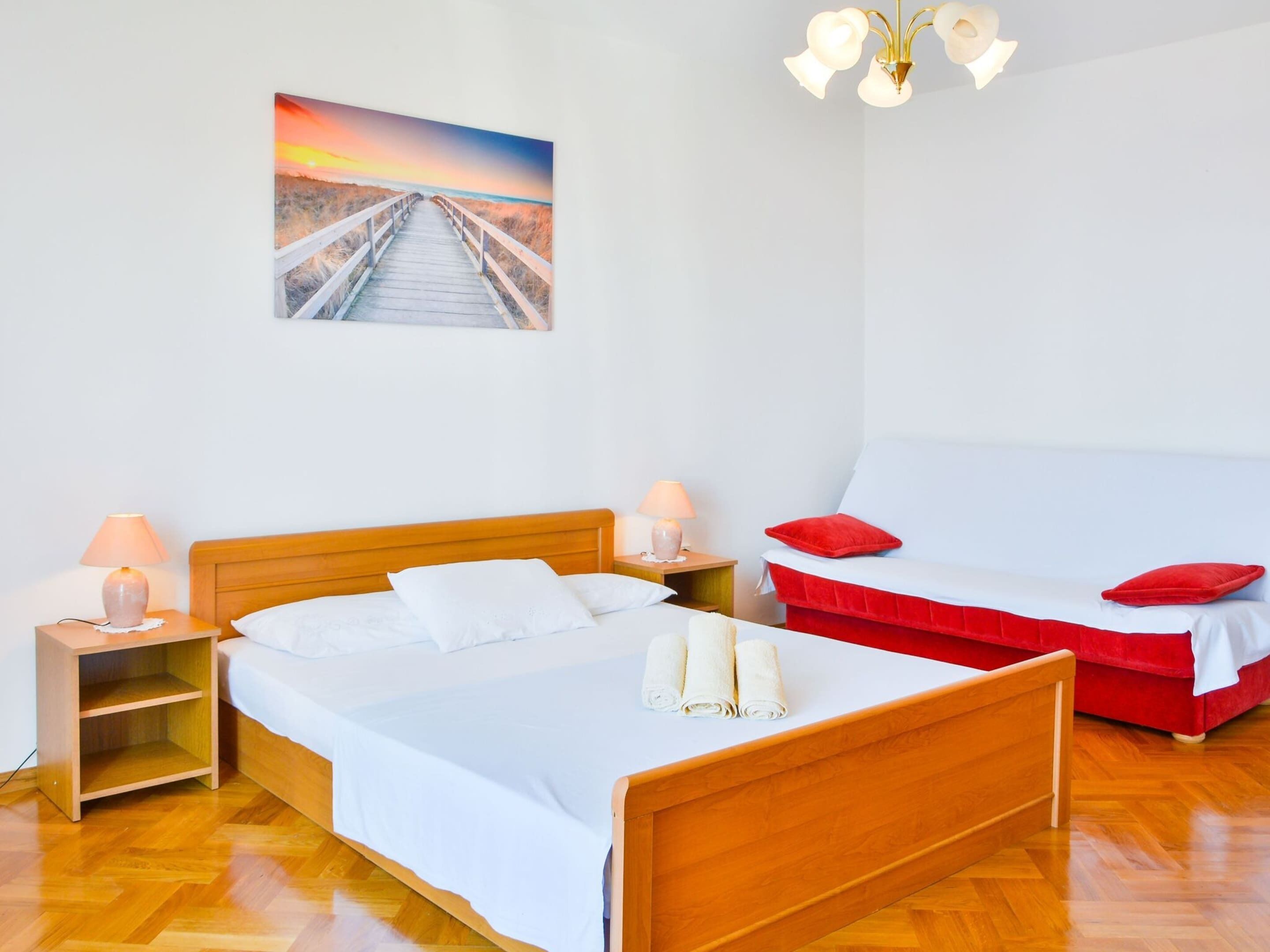 A bedroom with a wooden bed, a nightstand, a red sofa, a white wall, and a picture of a sunset over a beach.