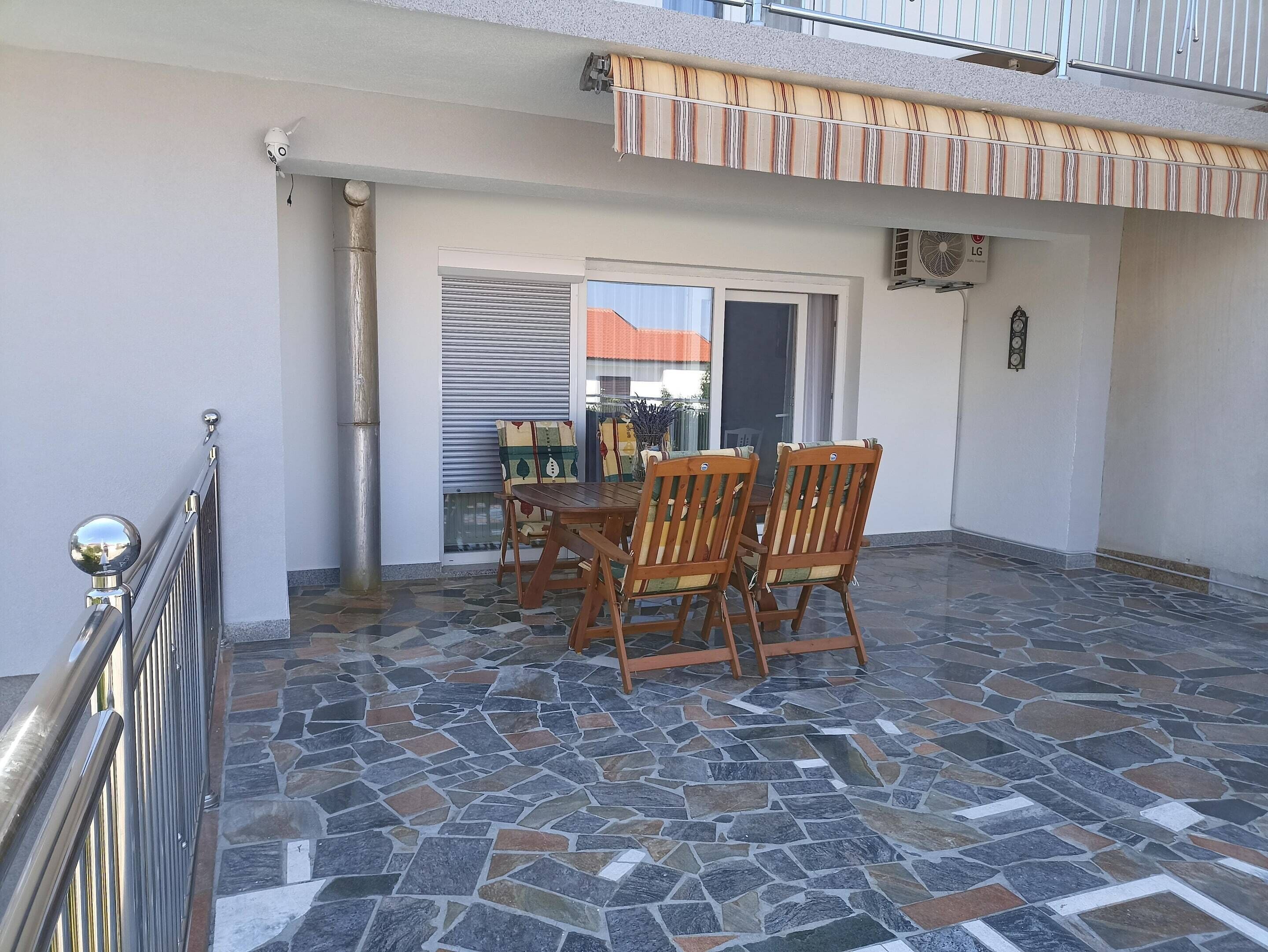 Une terrasse avec un sol en pierre, une table en bois entourée de chaises et une porte-fenêtre coulissante donnant sur un espace extérieur.