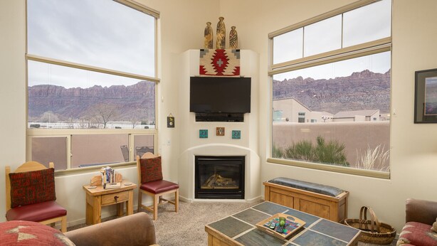 A cozy living room with a fireplace, large windows offering a view of a mountainous landscape, and a flat-screen TV mounted above the fireplace. - Great Location Near Parks | Pet-Friendly | Pool + Hot Tub Access (Moab)