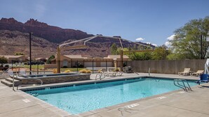 A rectangular pool with a clear blue water, surrounded by a concrete deck. There are lounge chairs and a small table on the deck. In the background, there are mountains and a clear sky. - Great Location Near Parks | Pet-Friendly | Pool + Hot Tub Access (Moab)