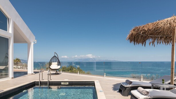 A poolside area with a view of the sea, a hanging chair, lounge chairs, and a thatched roof structure.