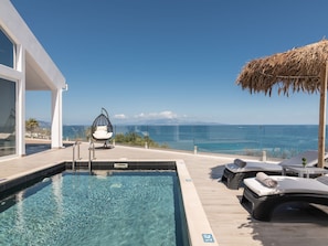 A poolside area with a view of the sea, a hanging chair, lounge chairs, and a thatched roof structure.