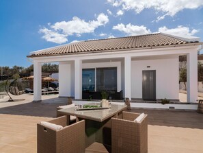 A modern outdoor patio with a glass table and wicker chairs, a covered seating area, and a pool in the background.