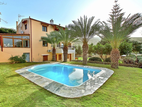 A modern two-story house with a swimming pool surrounded by a stone deck and lush greenery.