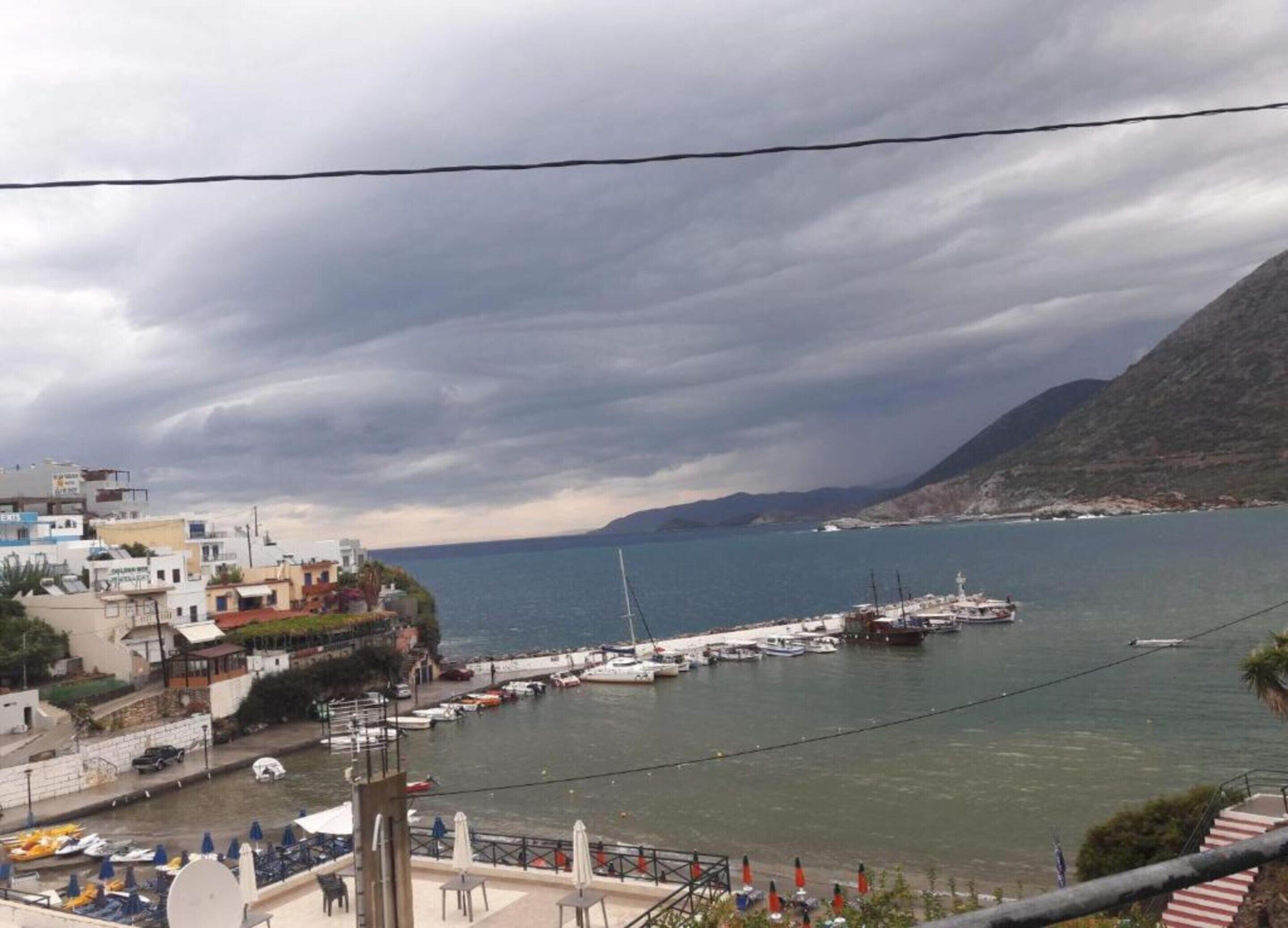 A coastal marina with boats docked, a mountainous backdrop, and buildings along the waterfront.