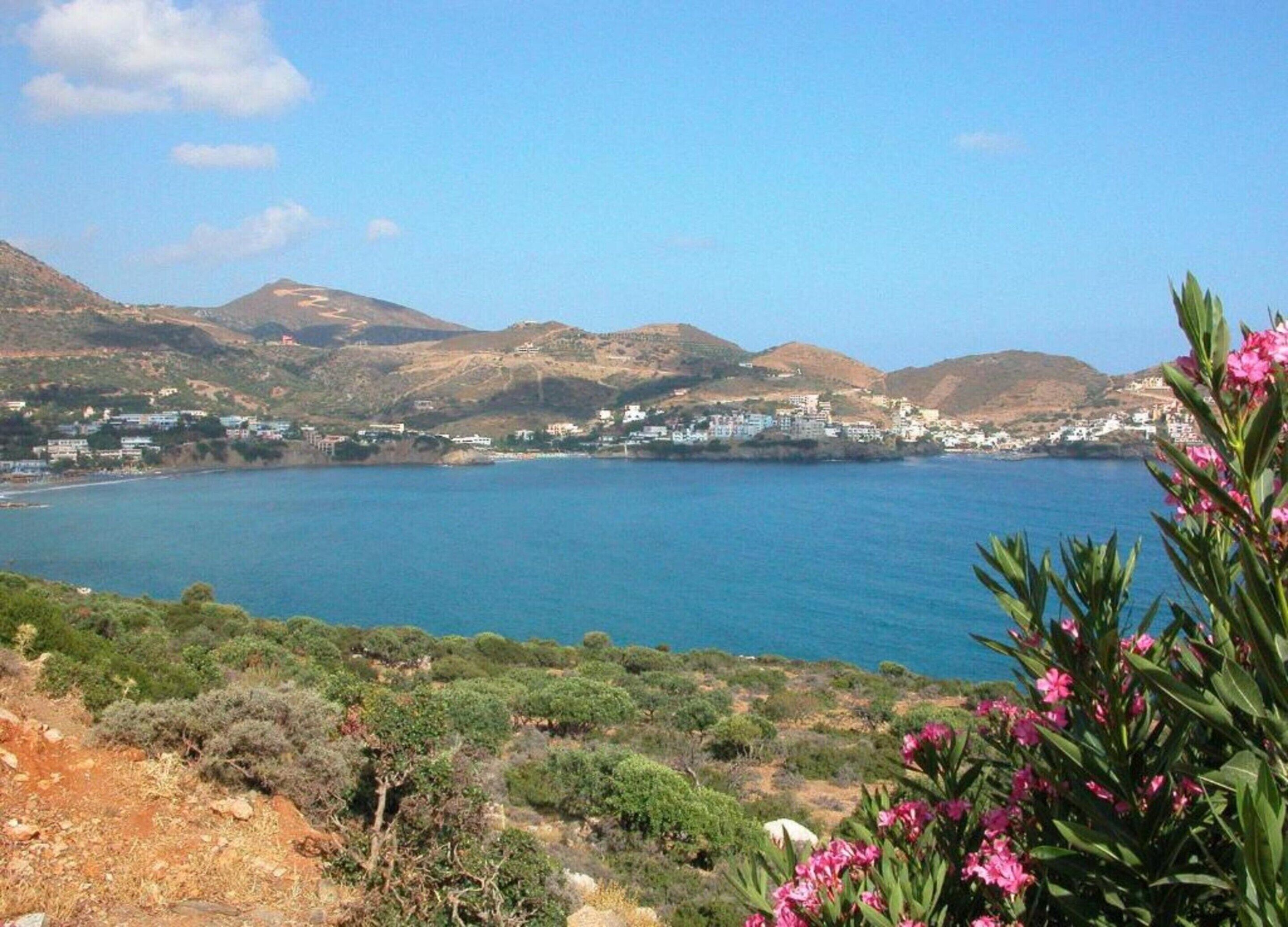 A coastal landscape with a clear view of the sea, hills, and a small settlement with buildings.