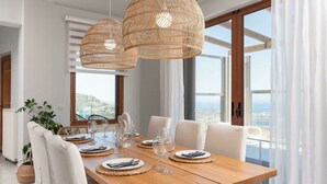 Uma área de refeições com uma mesa de madeira posta para quatro pessoas, cercada por cadeiras brancas. Há duas luminárias pendentes grandes e trançadas sobre a mesa.