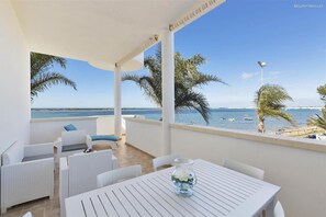 Een balkon met witte rieten meubels, een glazen vaas met bloemen en uitzicht op zee en palmbomen.