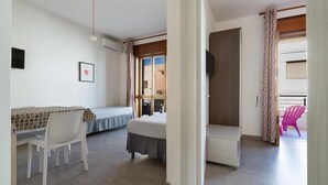 Una habitación compacta con cama, escritorio y silla. Cuenta con ventana con cortinas, puerta y un pequeño balcón con una silla.