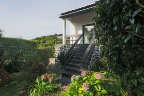 A modern house with a stone exterior, a staircase leading to a covered porch, and lush greenery surrounding the property. - O Nosso Refúgio (Água de Pau)